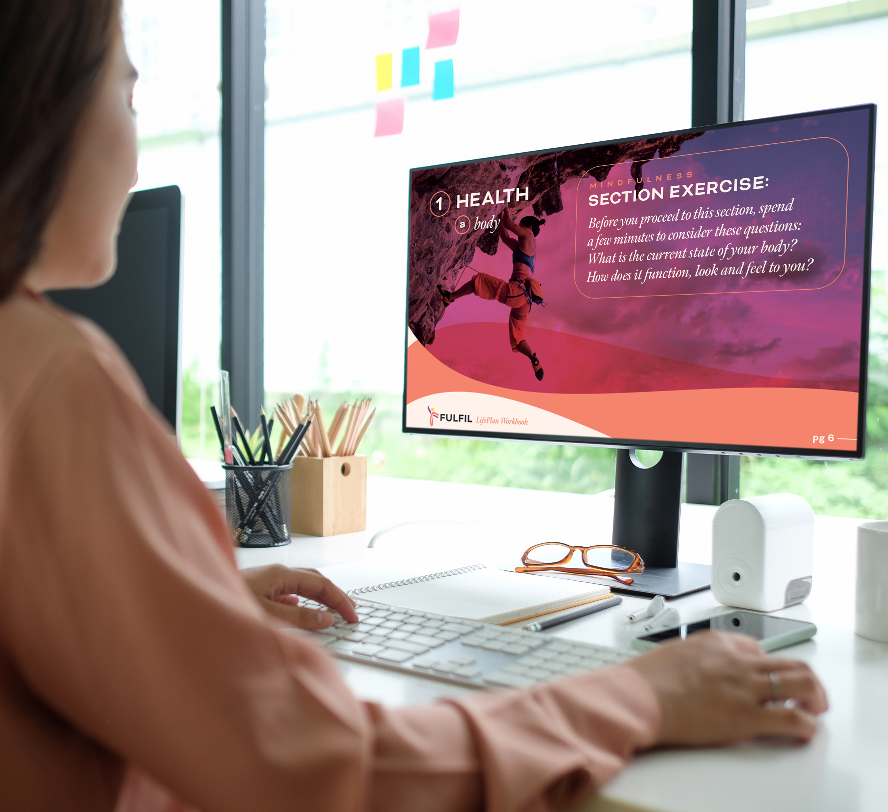 A woman wearing peach colored blous viewing the Fulfil LifePlan Workbook on her desktop computer in a natural light-filled office.