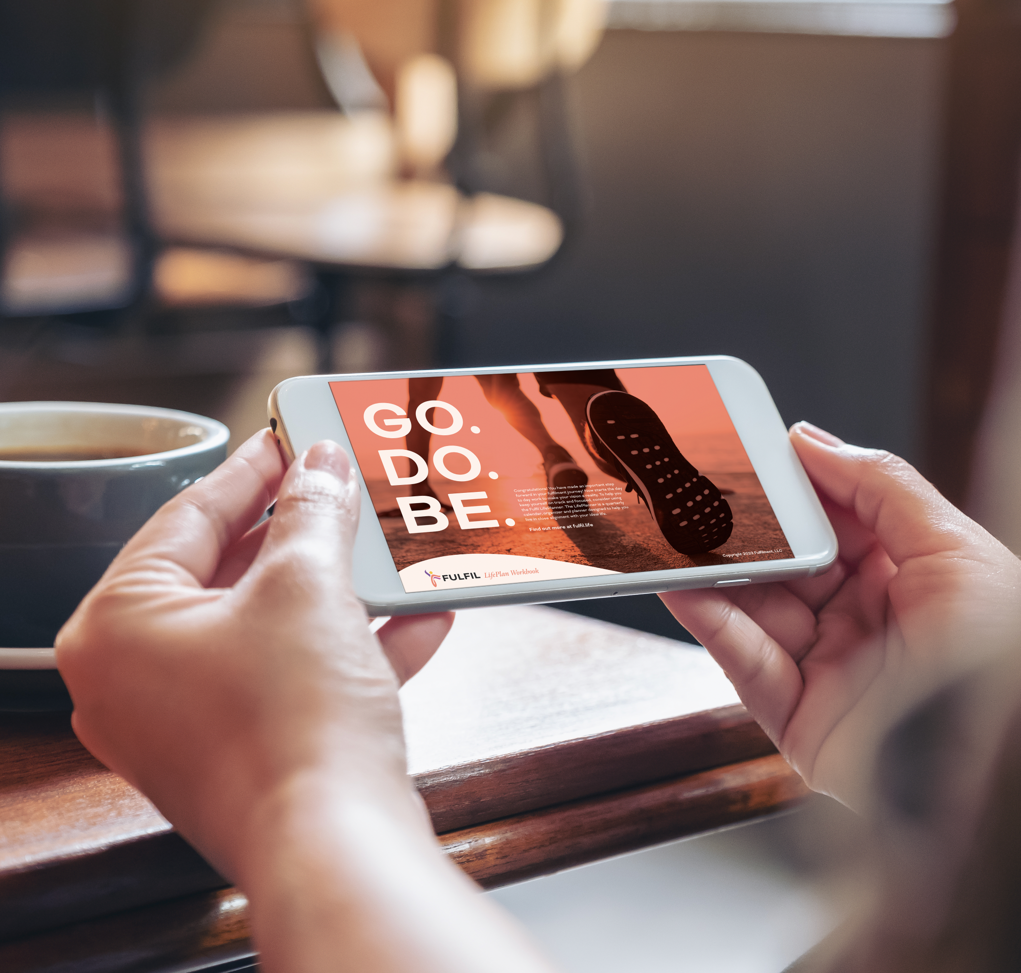 A persons hands holding a phone horizontally displaying the Fulfil LifePlan Workbook all in a coffee shop setting with a latte on the table.